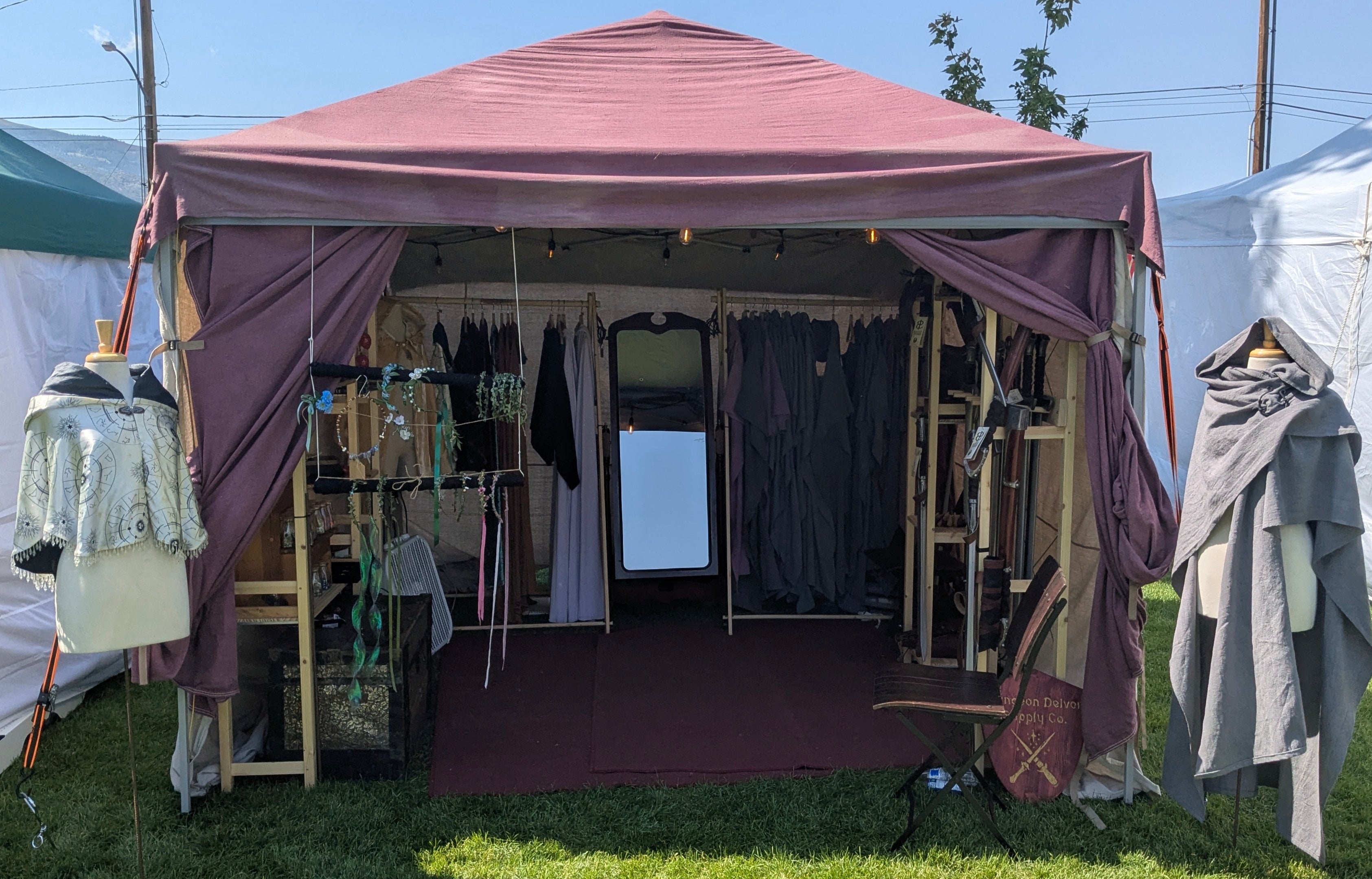 Outdoor Renaissance Faire style clothing tent with various garments, foam weapons, dice, and flower crowns on display under a clear blue sky.