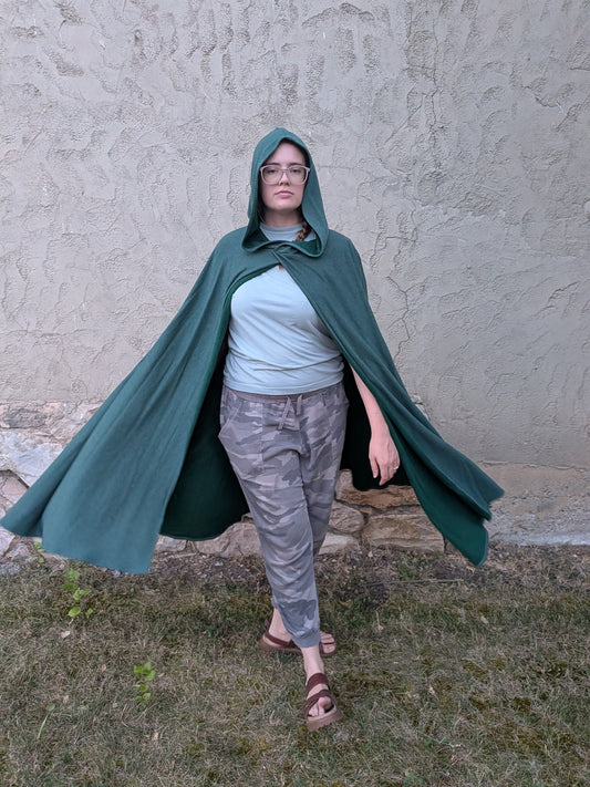 A person wearing a dark green hooded cape cloak, a light blue t-shirt, and gray camouflage pants, standing outdoors against a textured stucco wall.