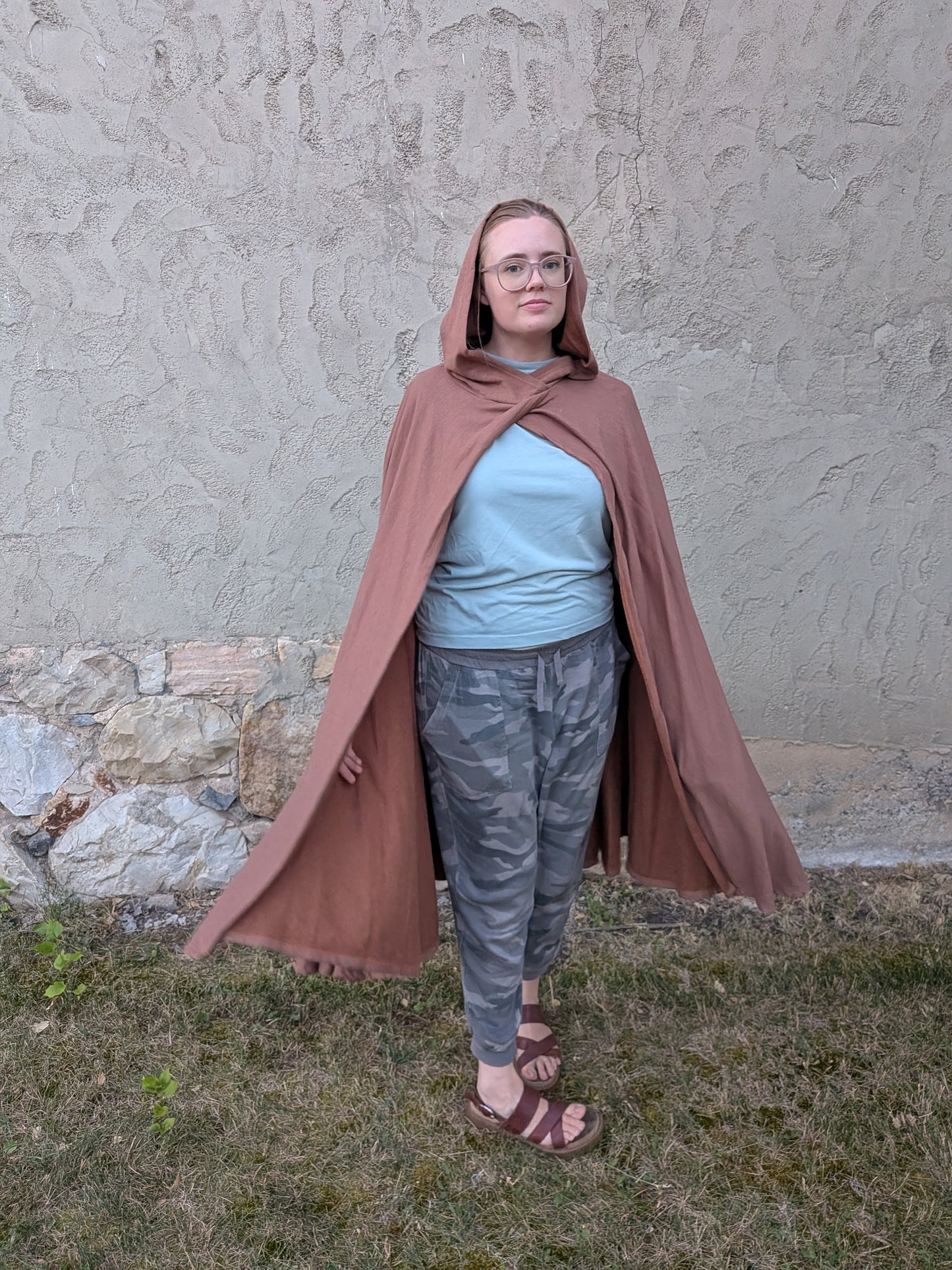 A person wearing a dusty rose-brown hooded cape stands outdoors against a textured stucco wall and grassy ground. The person is also wearing a light blue t-shirt, gray camouflage pants, and brown sandals.