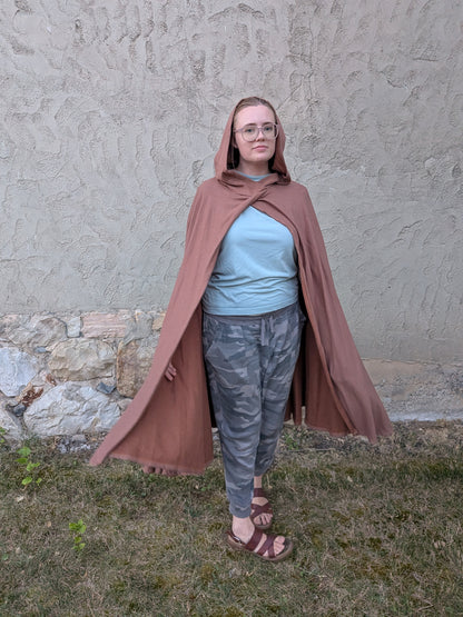 A person wearing a dusty rose-brown hooded cape stands outdoors against a textured stucco wall and grassy ground. The person is also wearing a light blue t-shirt, gray camouflage pants, and brown sandals.