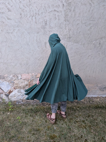 A person wearing a dark green hooded cloak or poncho, standing outdoors against a textured stucco wall and grassy ground.