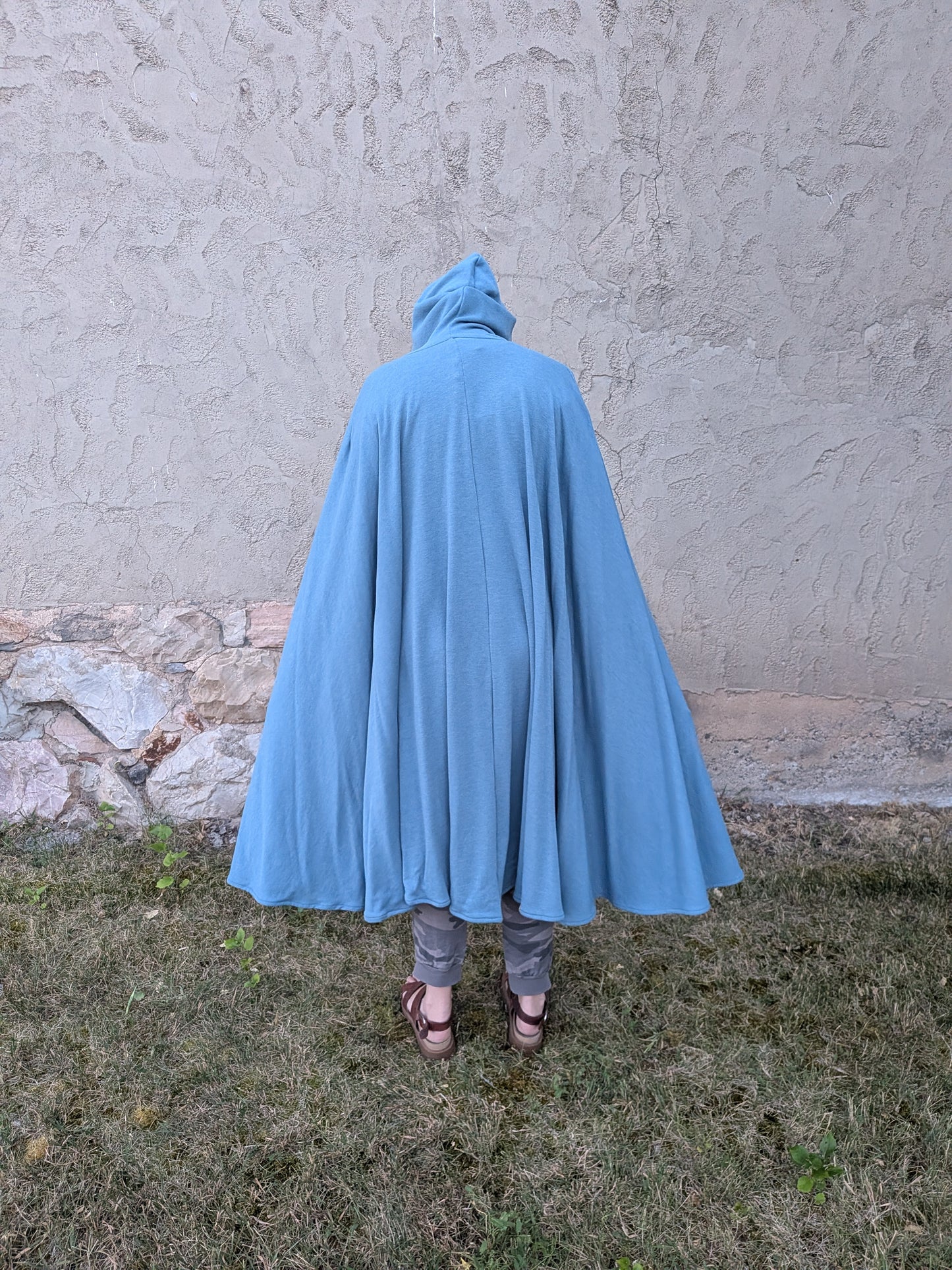 A person viewed from behind wearing a flowing light blue hooded cape or poncho, standing outdoors on grass in front of a textured stucco wall.