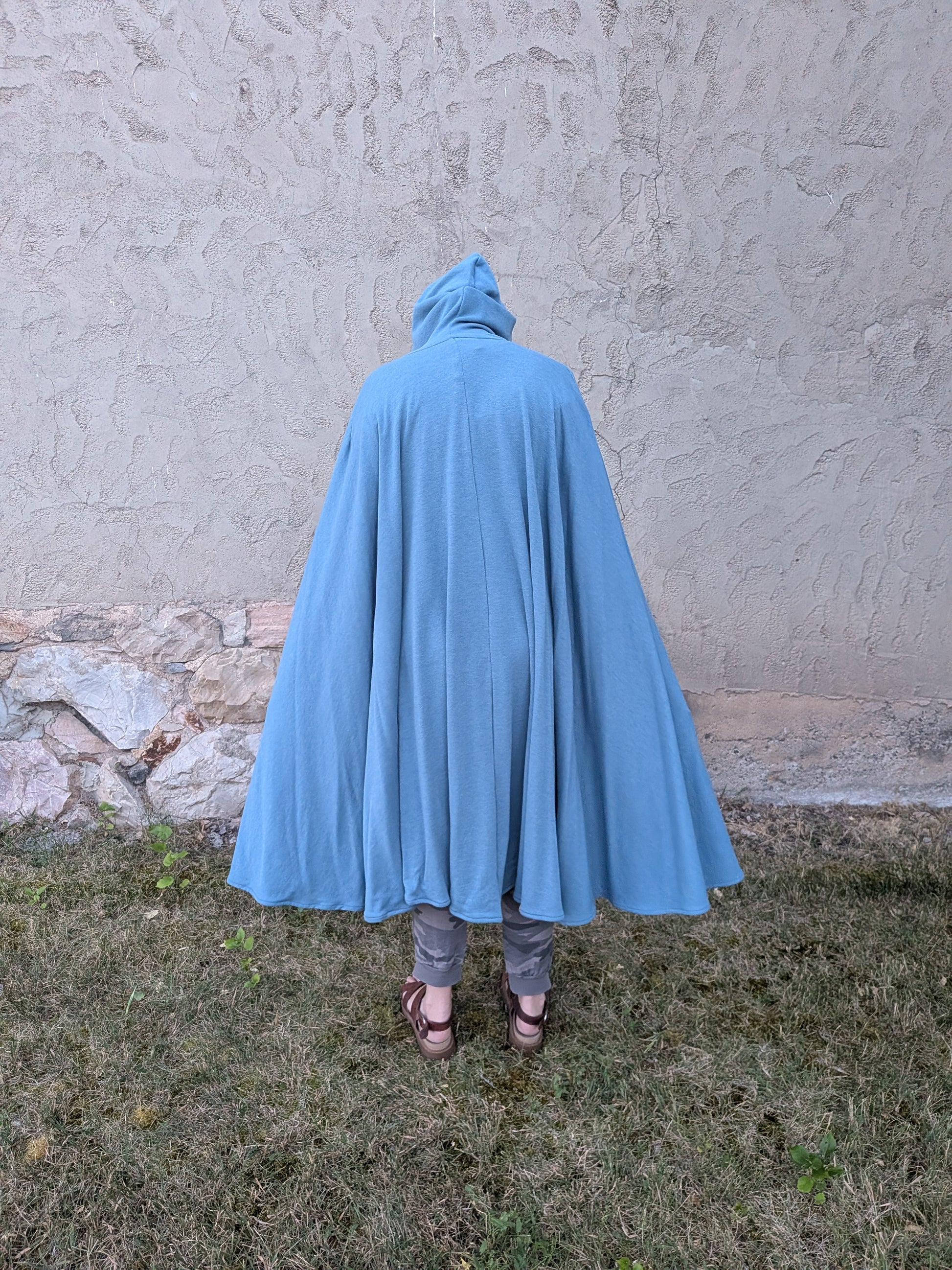 A person viewed from behind wearing a flowing light blue hooded cape or poncho, standing outdoors on grass in front of a textured stucco wall.