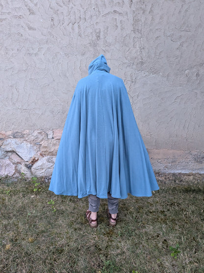 A person viewed from behind wearing a flowing light blue hooded cape or poncho, standing outdoors on grass in front of a textured stucco wall.