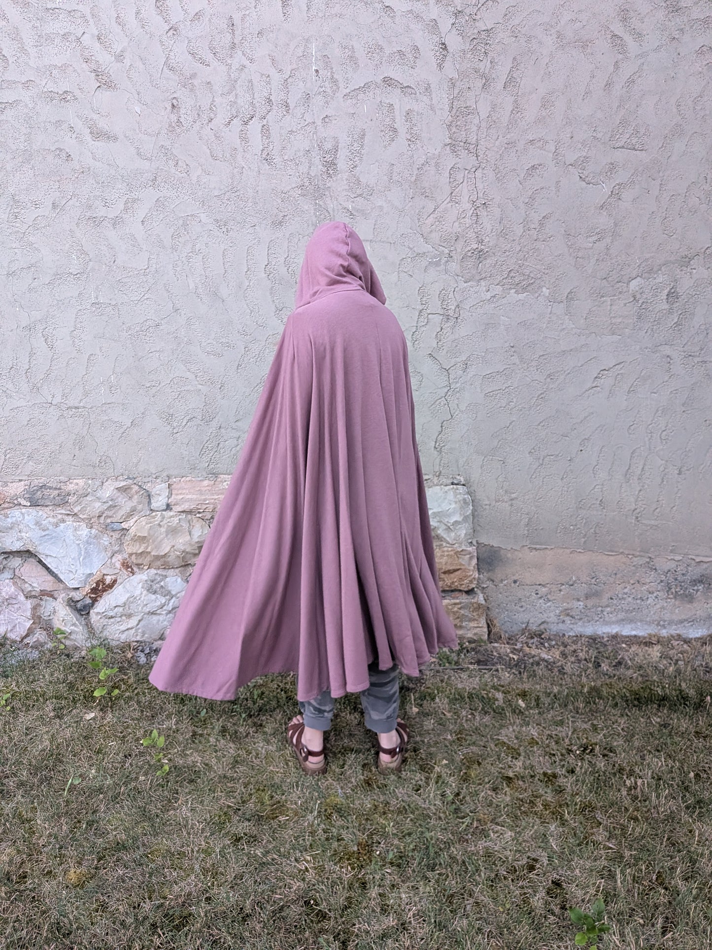 A person viewed from behind wearing a dusty mauve-purple hooded cape or poncho, standing outdoors on grass against a textured stucco wall.