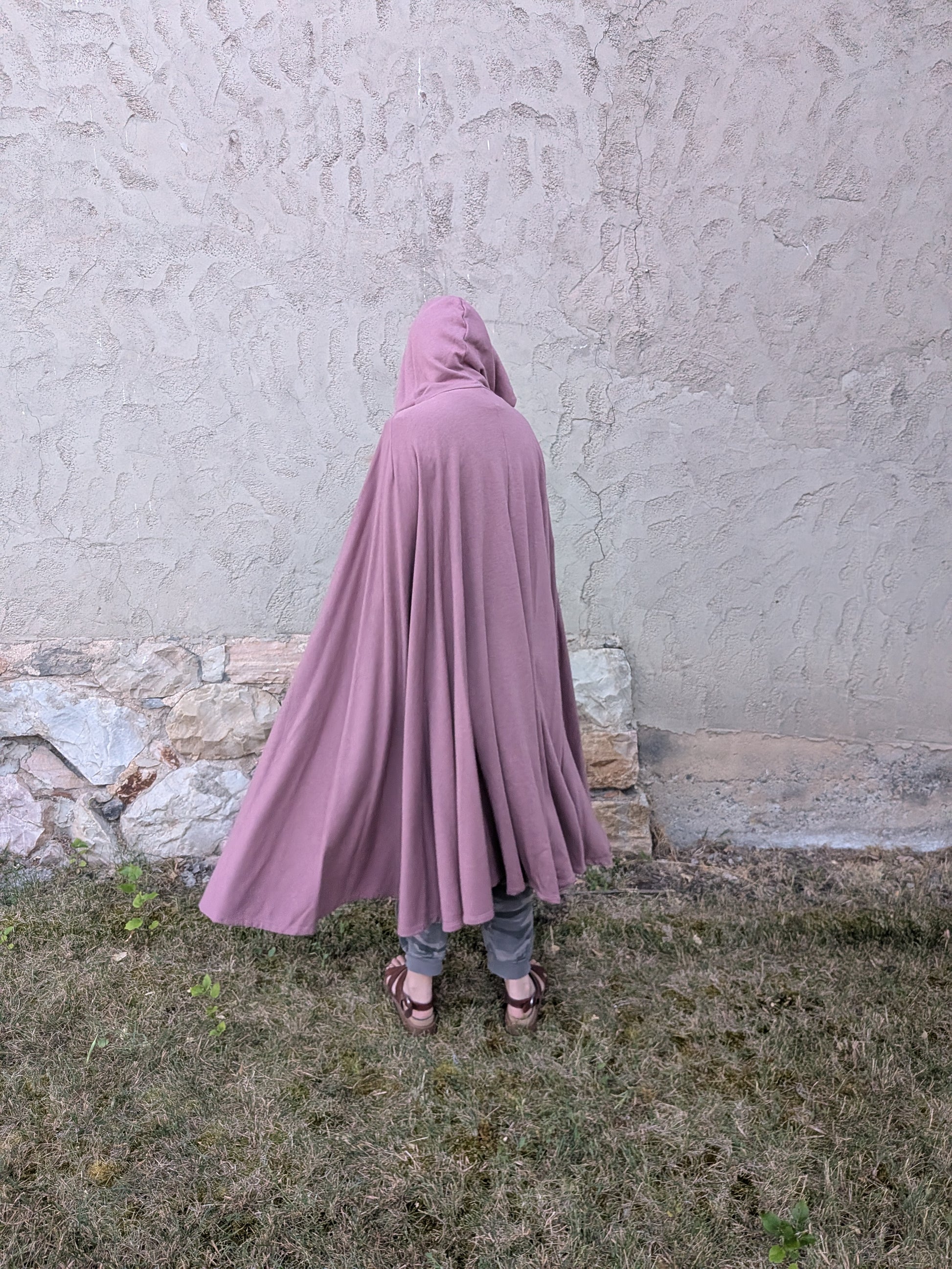 A person viewed from behind wearing a dusty mauve-purple hooded cape or poncho, standing outdoors on grass against a textured stucco wall.