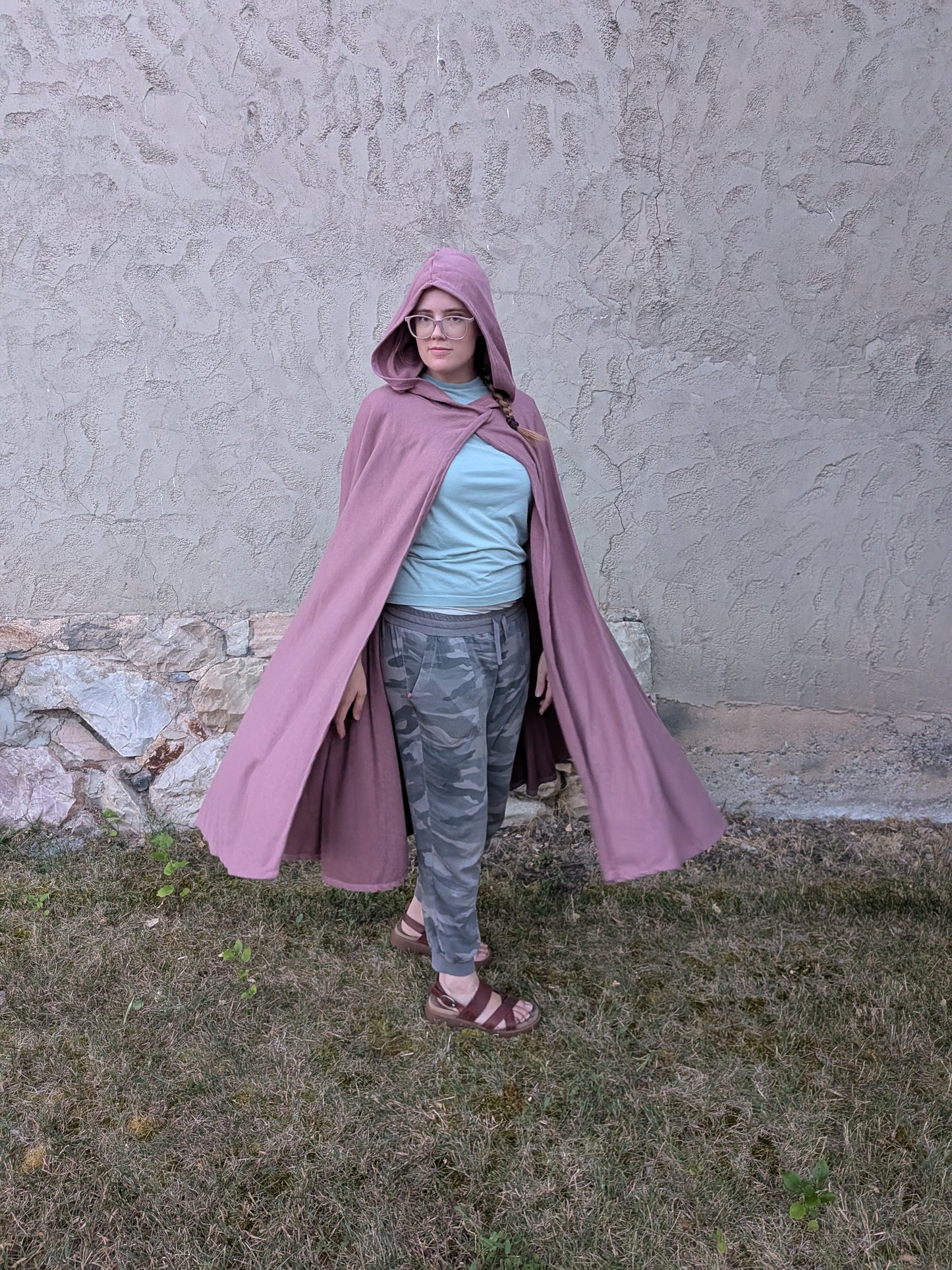 A person wearing a dusty pink hooded cape over a light blue t-shirt, camouflage pants, and brown sandals, standing outdoors against a textured stucco wall.