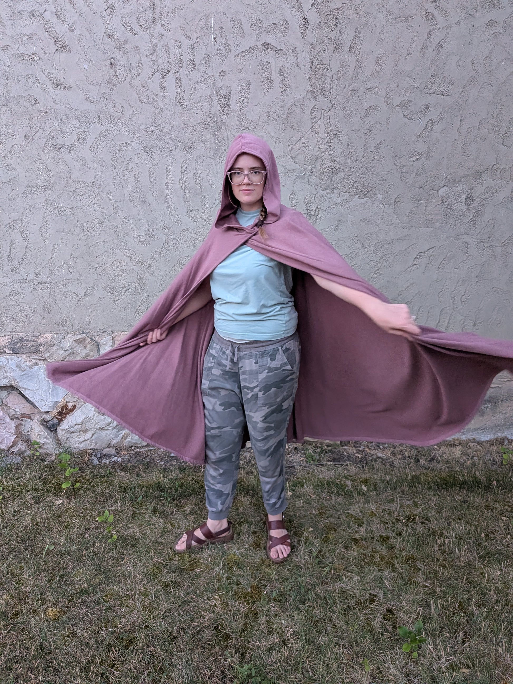 A person wearing a dusty purple hooded cape stands outdoors on grass in front of a textured stucco wall, holding the cape out to both sides.