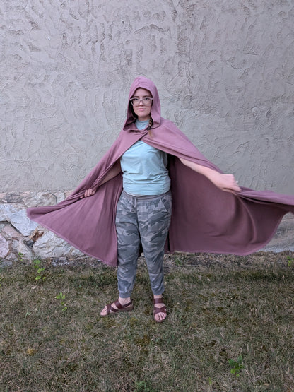 A person wearing a dusty purple hooded cape stands outdoors on grass in front of a textured stucco wall, holding the cape out to both sides.