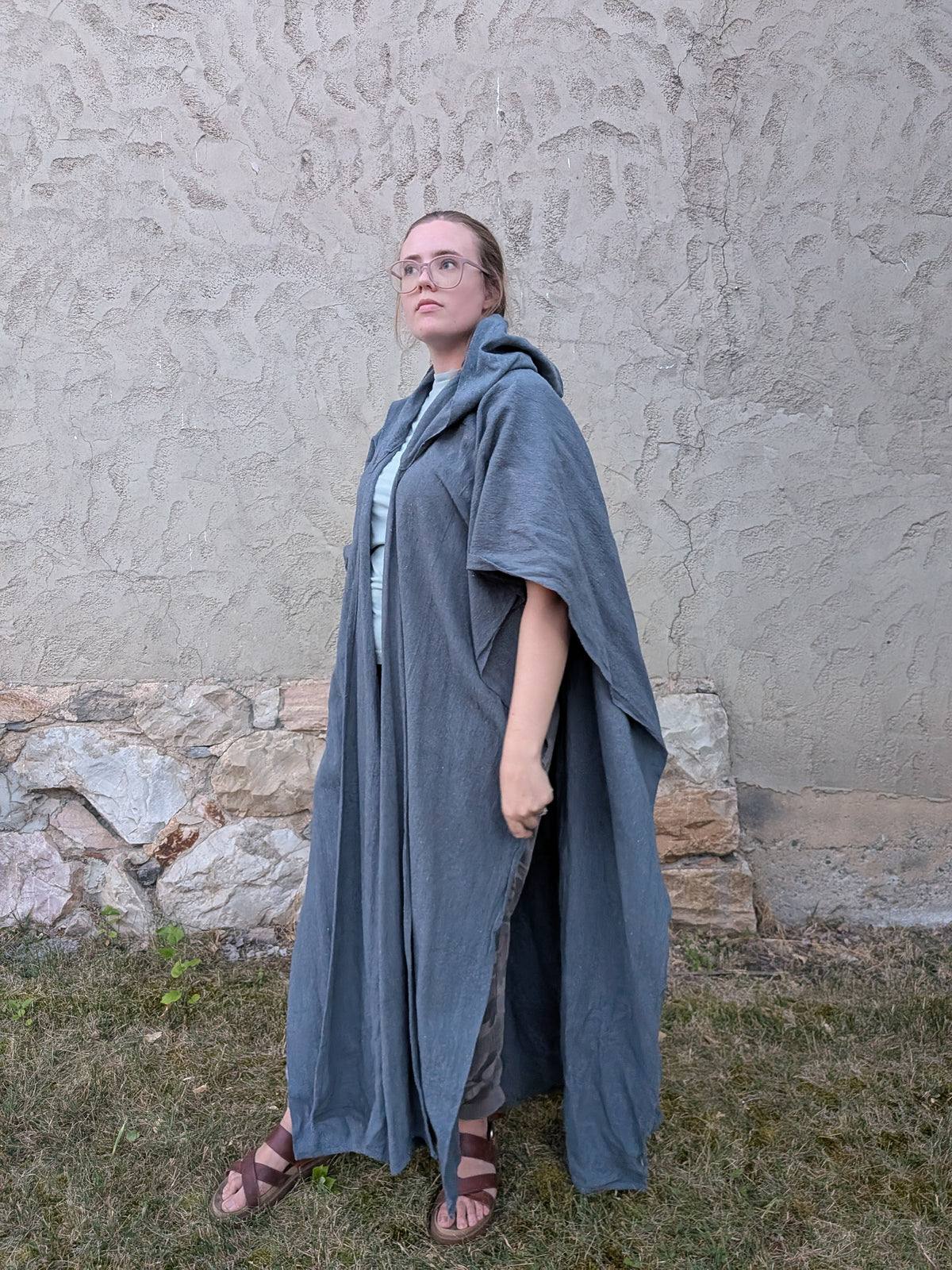 A woman wearing a long, flowing slate blue linen robe or cloak over a light blue top and brown sandals, standing outdoors against a textured stucco wall and grass.