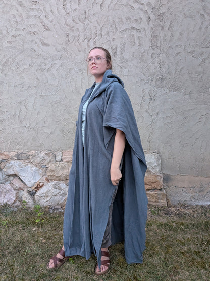 A woman wearing a long, flowing slate blue linen robe or cloak over a light blue top and brown sandals, standing outdoors against a textured stucco wall and grass.