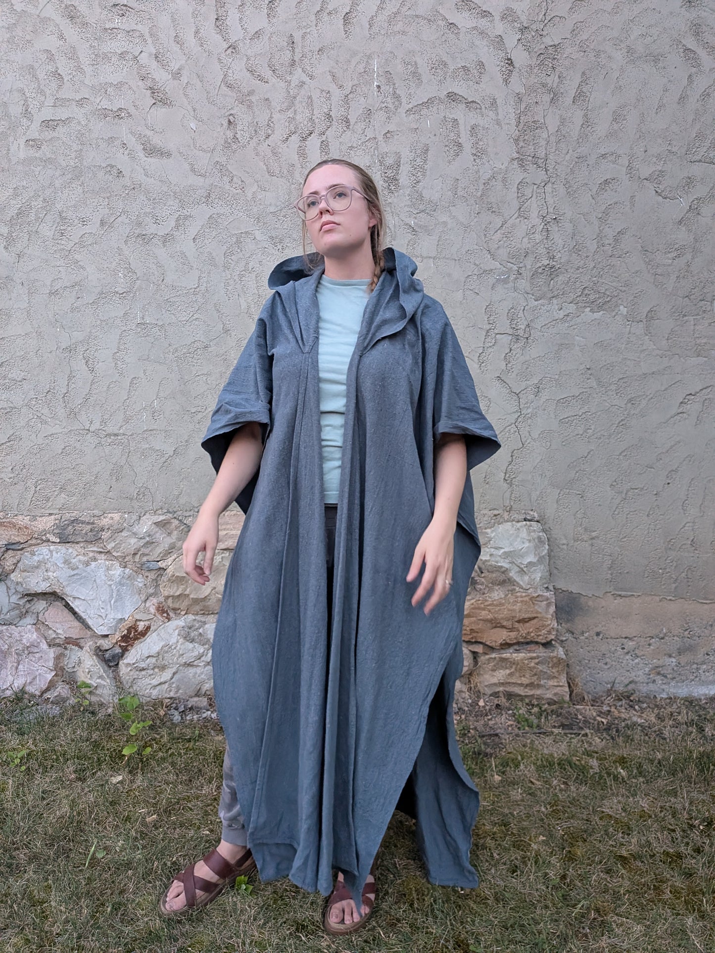 A woman wearing a long, loose-fitting slate blue-gray hooded poncho over a light blue top and brown sandals, standing outdoors against a textured stucco wall and grass.