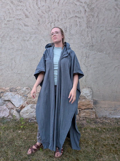 A woman wearing a long, loose-fitting slate blue-gray hooded poncho over a light blue top and brown sandals, standing outdoors against a textured stucco wall and grass.