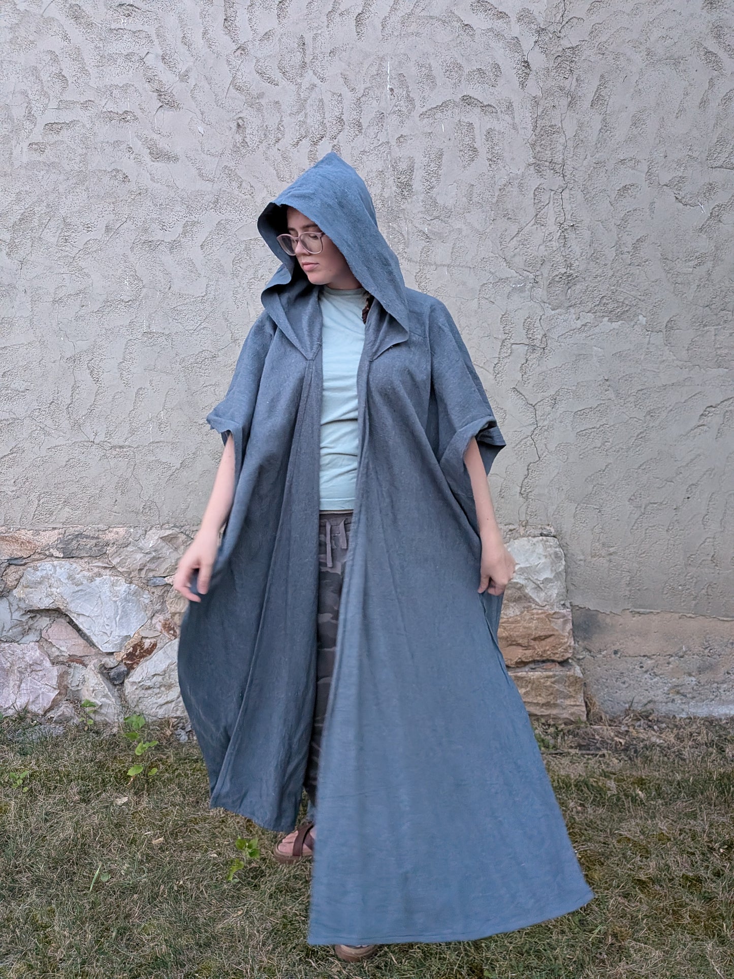 A person wearing a long, hooded blue-gray cloak or poncho over a light blue top and patterned pants, standing outdoors against a textured stucco wall and grass.
