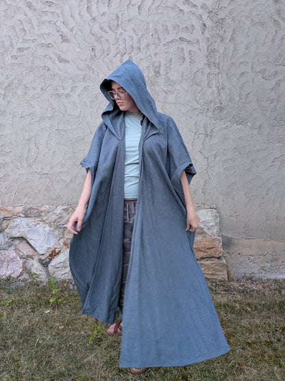 A person wearing a long, hooded blue-gray cloak or poncho over a light blue top and patterned pants, standing outdoors against a textured stucco wall and grass.