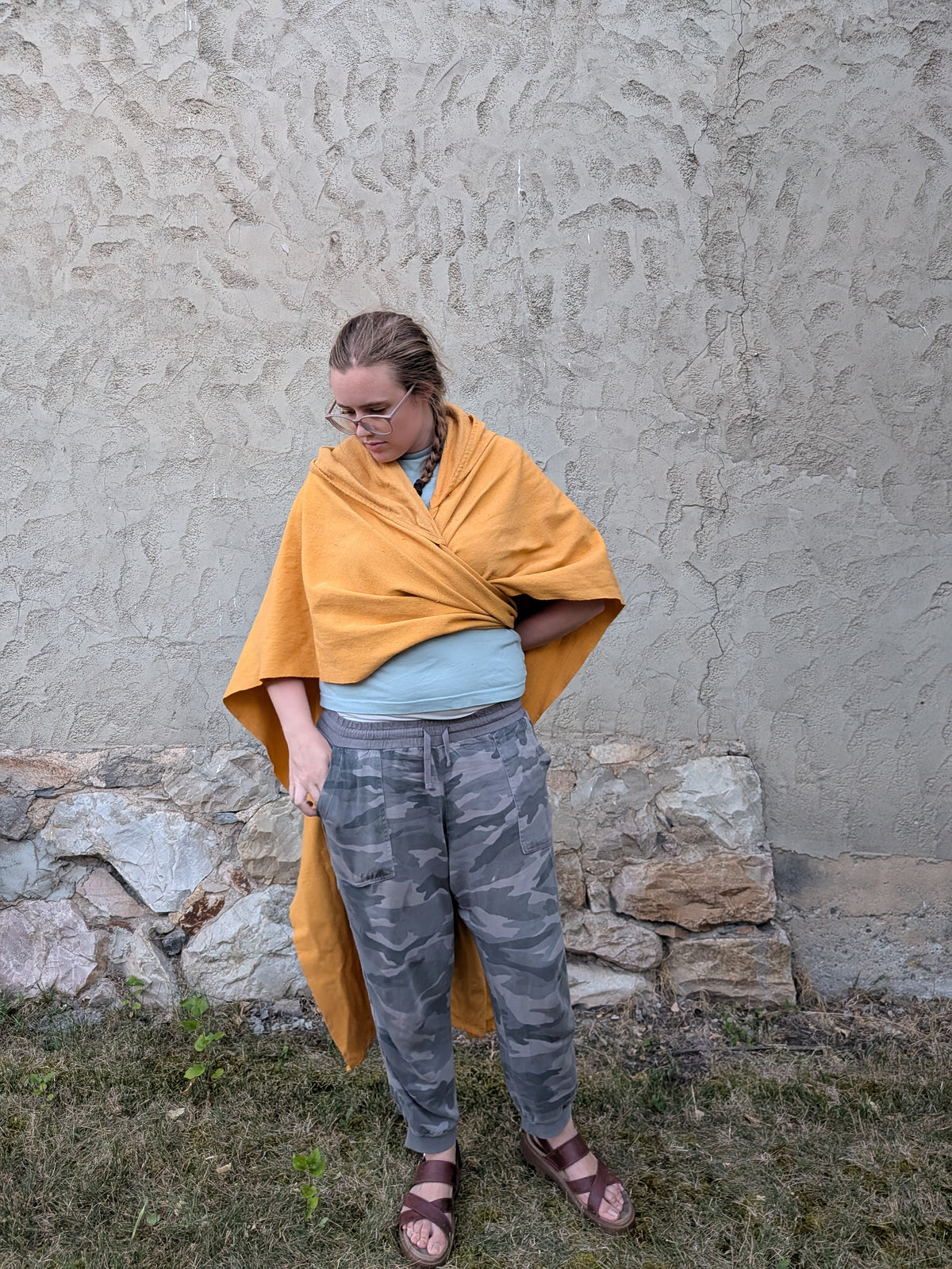 A person standing outdoors in front of a textured stucco wall and stone base, wearing a mustard yellow wrap or poncho draped over a light blue top and gray camouflage pants.