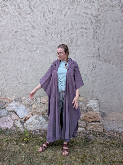 A woman wearing a loose, draped purple linen kimono-style cover-up over a light blue t-shirt and jeans, standing outdoors against a textured stucco wall.
