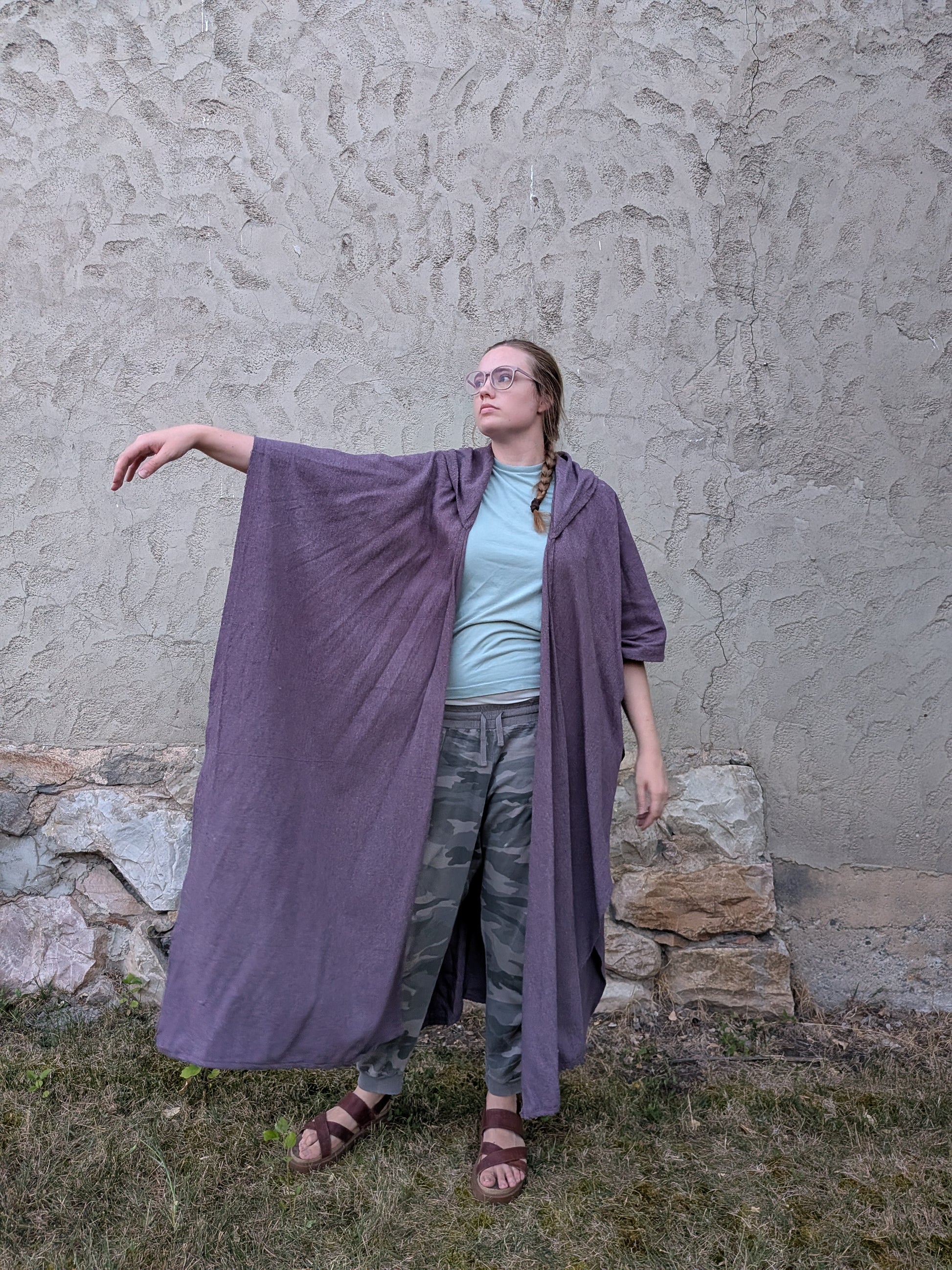 A woman wearing a long, flowing mauve-purple linen cloak or poncho over a light blue t-shirt, camouflage pants, and sandals, standing outdoors against a textured gray stucco wall.