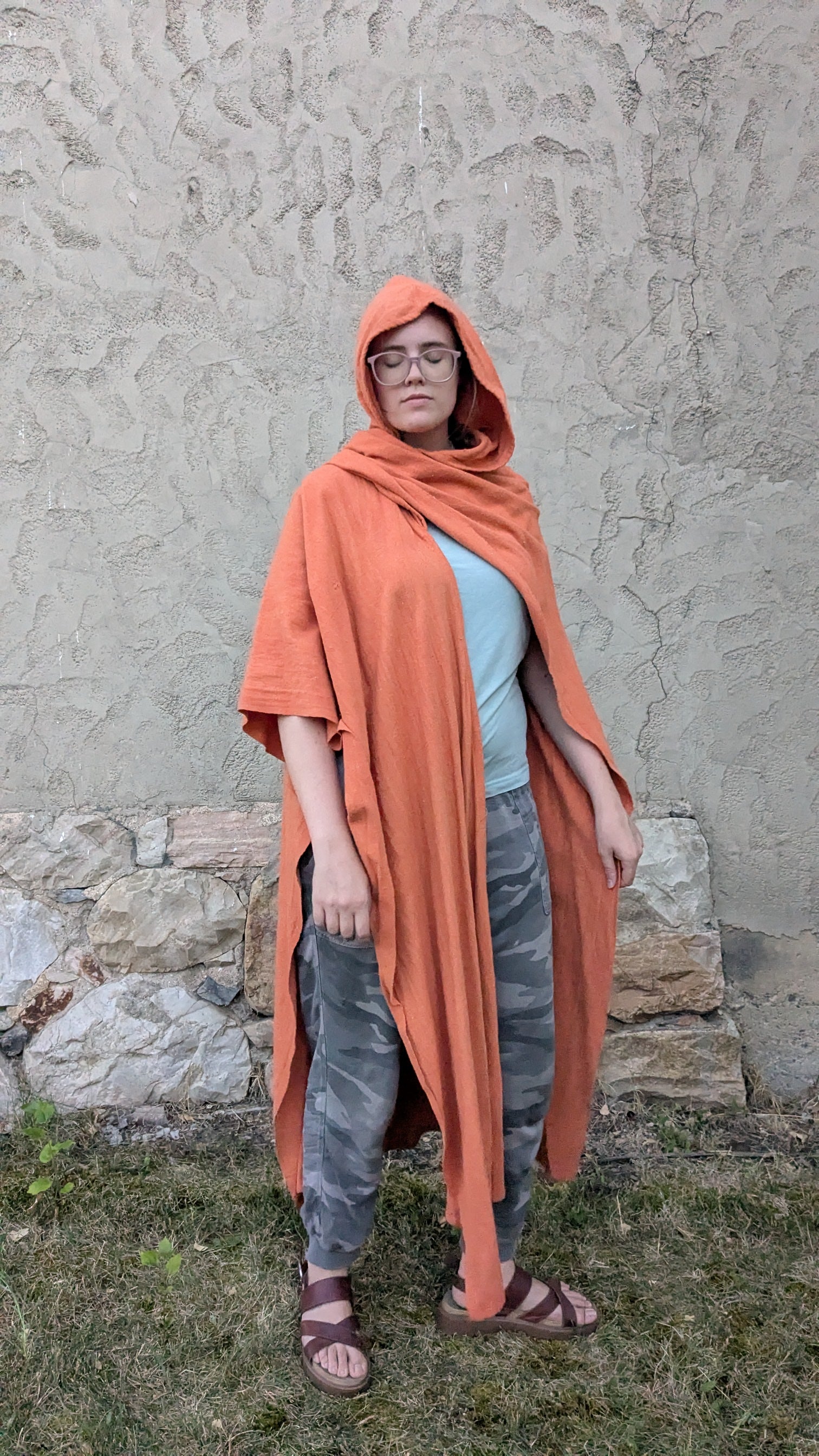A person wearing a bright orange hooded shawl or poncho over a light blue top and gray patterned pants, standing outdoors against a textured stucco wall and grass.