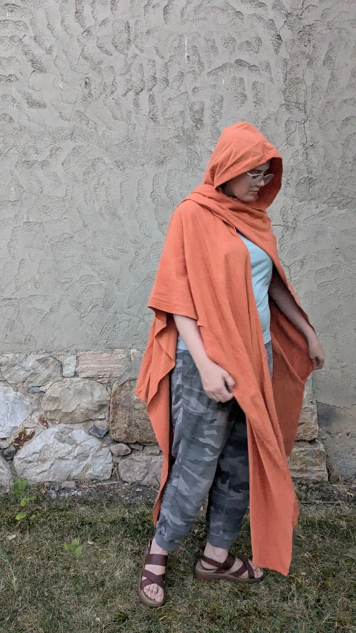 A person wearing a terracotta orange hooded poncho or cape over a light blue top and gray camouflage pants, standing outdoors against a textured stucco wall and grass.