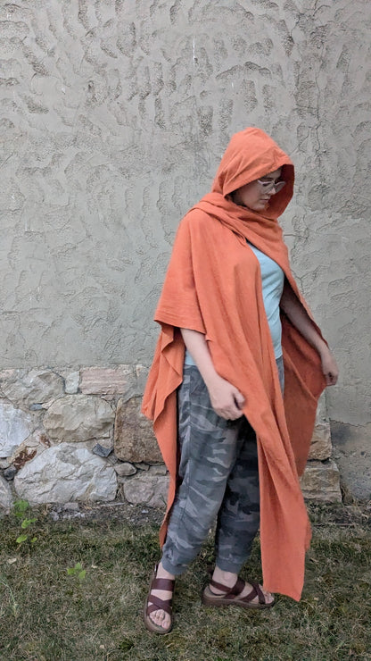 A person wearing a terracotta orange hooded poncho or cape over a light blue top and gray camouflage pants, standing outdoors against a textured stucco wall and grass.