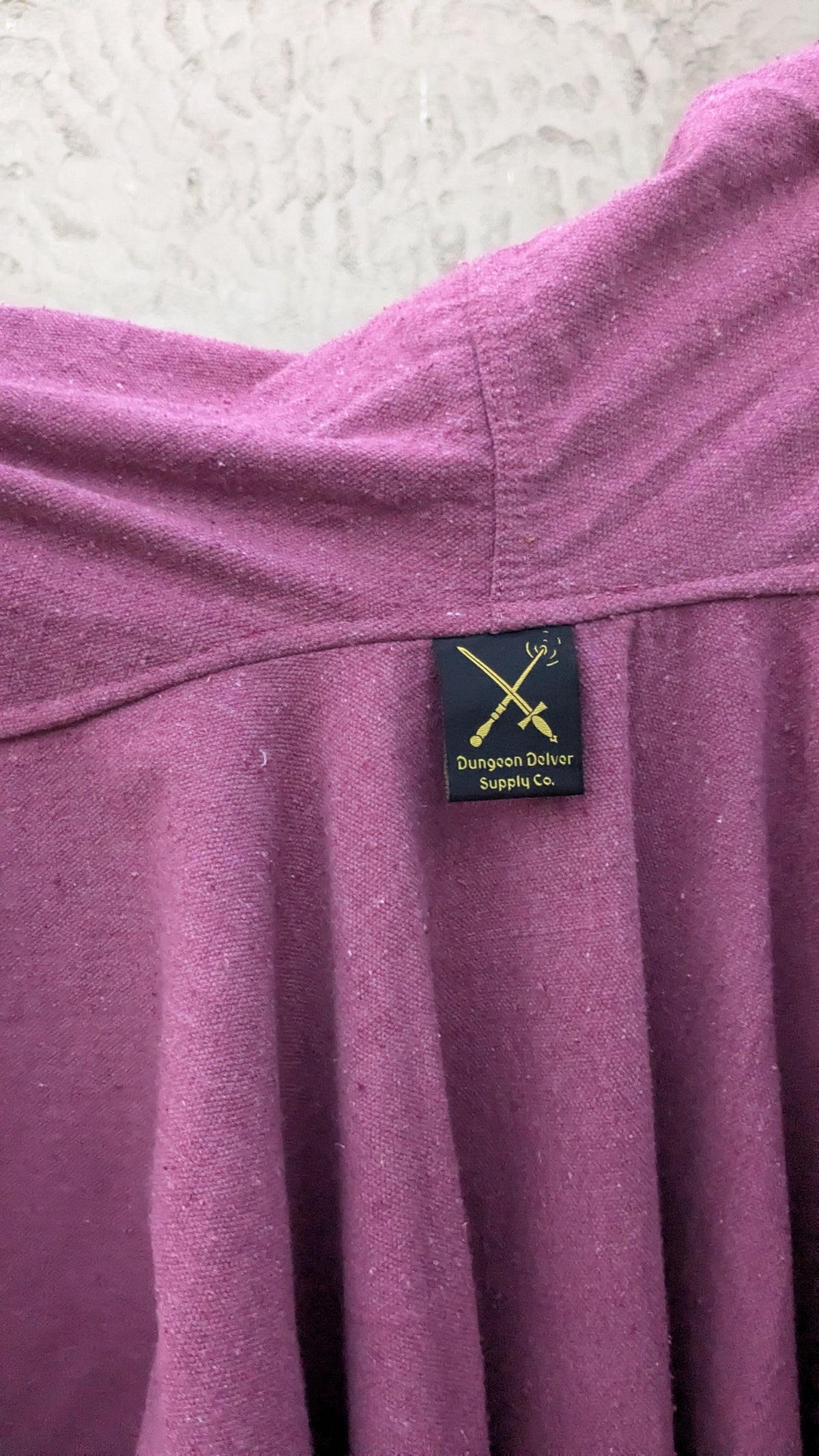 Close-up of a dusty purple or mauve knit garment, likely a dress, featuring a black sewn-in label with gold text and logo that reads 'Dungeon Delver Supply Co.'.