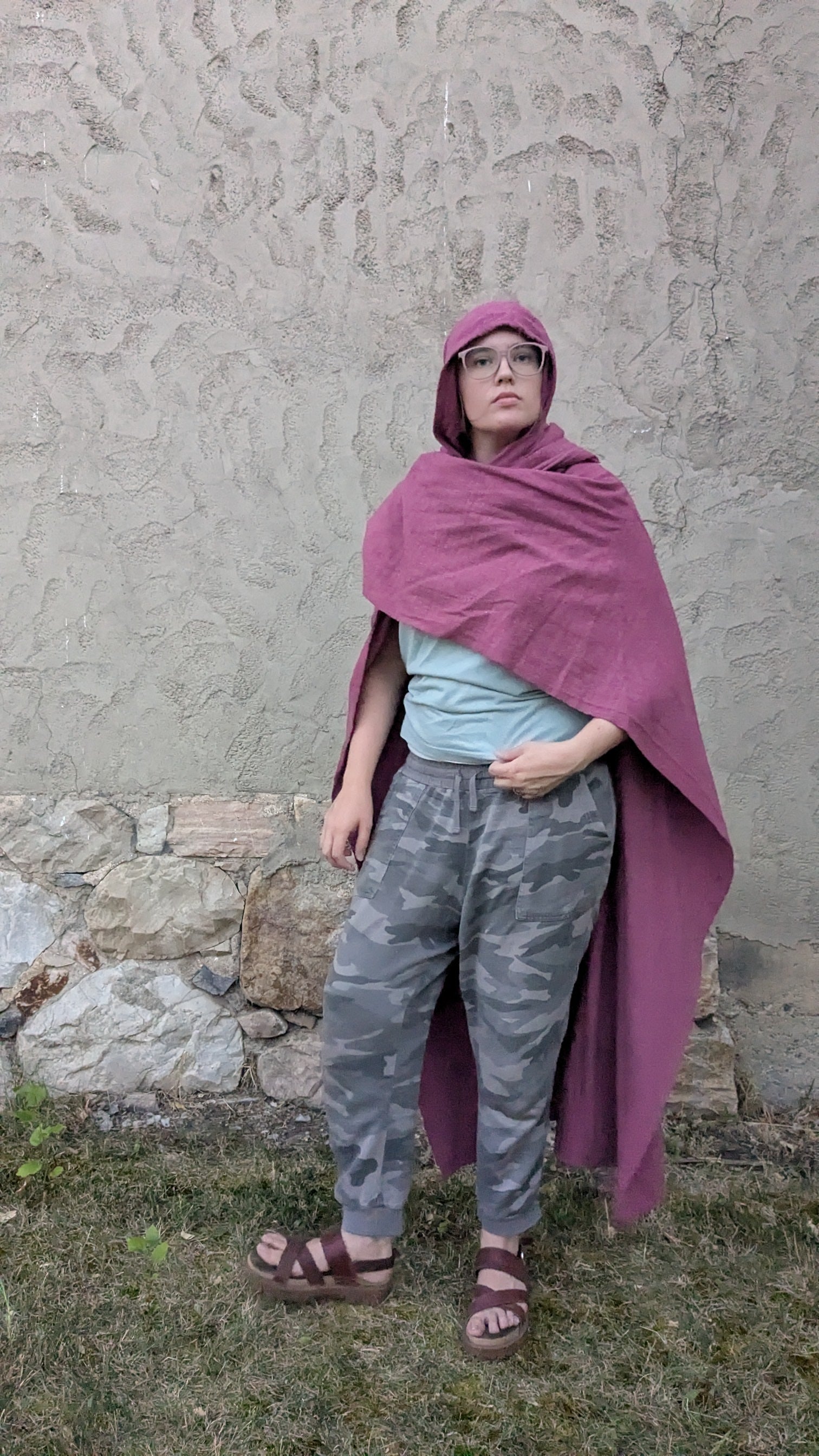 A person wearing a dusty purple hooded cape or poncho over a light blue t-shirt, gray camouflage pants, and brown sandals, standing outdoors against a textured stucco wall.