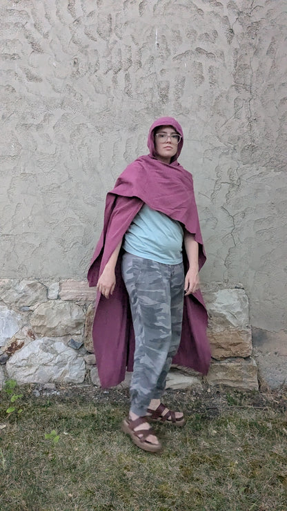 A person wearing a dusty purple hooded cloak over a light blue t-shirt, gray camouflage pants, and brown sandals, standing outdoors against a textured stucco wall.